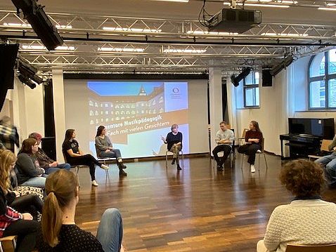 Gruppe sitzt im Stuhlkreis während der Studiengang Elementare Musikpädagogik vorgestellt wird. Präsentation mit dem Titel des Studiengangs wird hinten an der Wand des Raumes gezeigt. 
