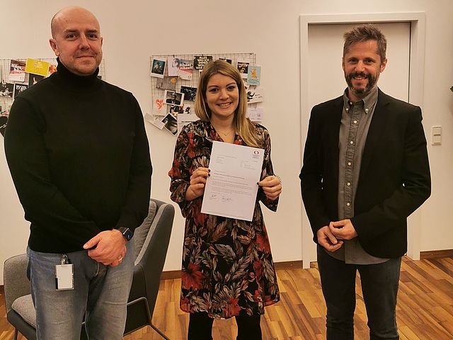 Kanzler Alexander Würth (li.) und Hochschulpräsident Prof. Rainer Kotzian (re.) gratulieren Marina Weber. Sie steht in der Mitte und hält den Brief in den Händen. 