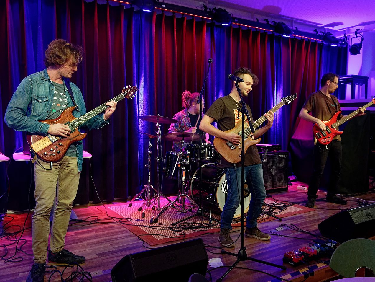Vier Herren stehen auf der Bühne der Cafeteria. Drei von ihnen stehen in einer Reihe und spielen Gitarre/Bass, hinter ihnen sitzt ein Mann am Schlagzeug. Die Wand im Hintergrund ist blau und rot angeleuchtet. 