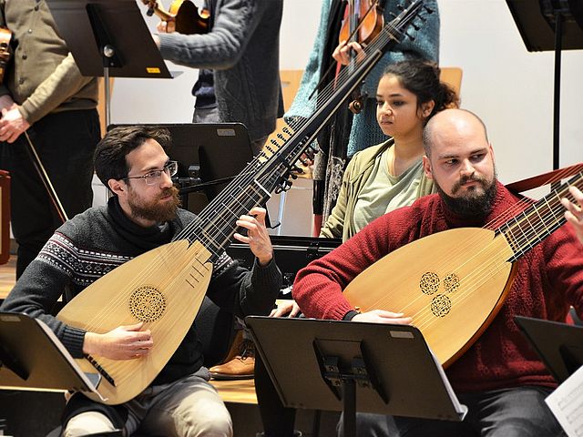 Zwei Männer sitzen mit ihren Lauten nebeneinander. Im Hintergrund befinden sich weitere Musiker*innen mit ihren Instrumenten. 