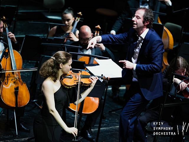 Im Vordergrund befindet sich die Solistin, Violine spielend. Im Hintergrund sind Dirigent und Orchester zu sehen, rechts unten im Bild befindet sich das Logo des Kyiv Symphony Orchestra. 