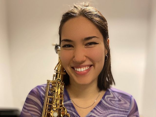 Porträt der jungen Frau mit kurzen, dunklen Haaren. In der rechten Hand hält sie ihr Saxophon. 