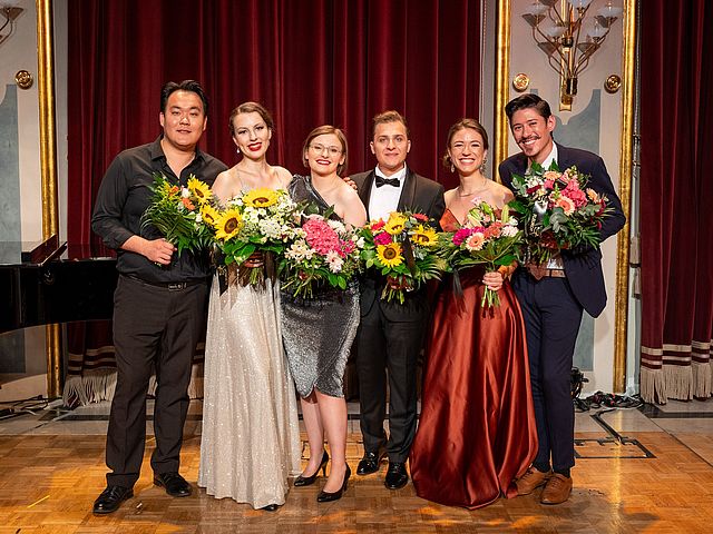 Die sechs Musiker*innen stehen lächelnd in einer Reihe und halten Blumensträuße in den Händen.