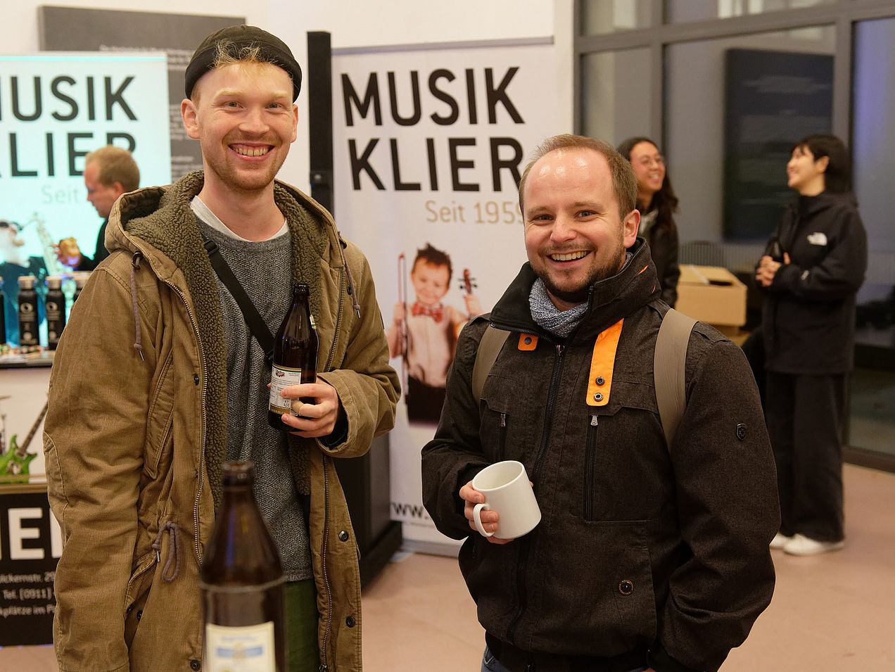 Zwei Studenten stehen lächelnd nebeneinander im Foyer der Hochschule mit Bier bzw. einer Tasse in der Hand