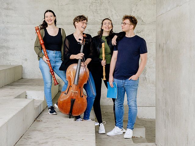 Die vier Mitglieder des Ensembles (Tabea Wink und Marie Erndl (Blockflöten), Anna Maria Rudolph (Cello) und Dominik Heidl (Cembalo)) stehen nebeneinander mit ihren Instrumenten auf Beton-Stufen und lachen.