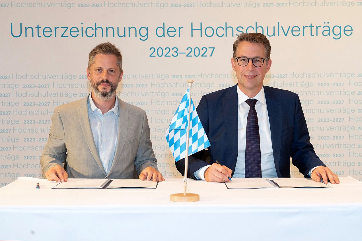 Prof. Rainer Kotzian und Markus Blume sitzen an einem Tisch. Zwischen ihnen steht eine kleine bayrische Flagge, vor ihnen liegen die unterzeichneten Verträge. 