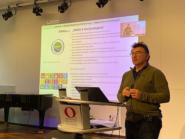 Dr. Frank Zirkl steht am Redner-Pult, hinter ihm ist seine Präsentation zu EMAS und Nachhaltigkeit an die Wand projeziert. 
