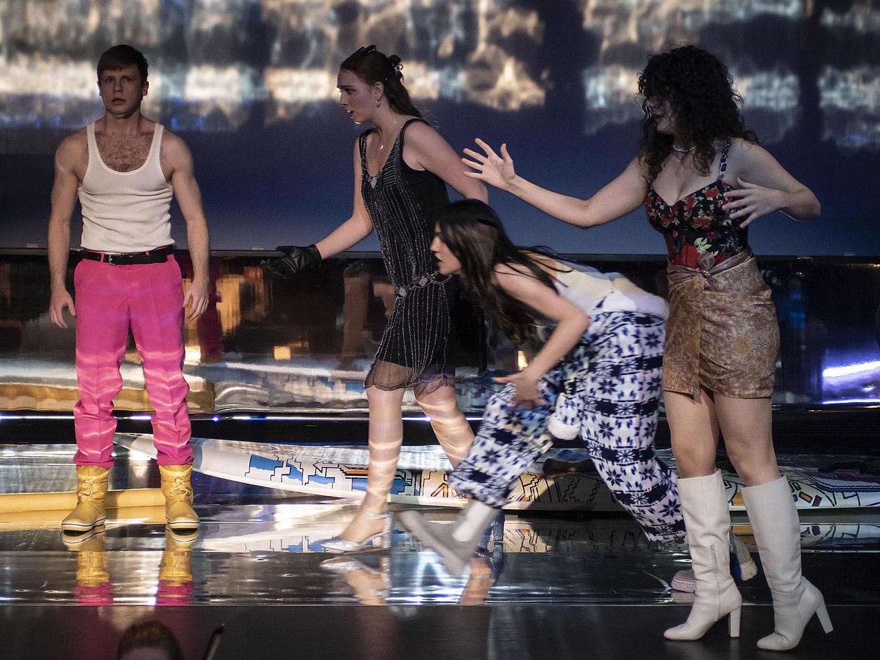 Links im Bild steht ein Darsteller mit sehr angespannter Körperhaltung. Rechts von ihm läuft eine Darstellerin quer über die Bühne, eine weitere Darstellerin folgt ihr in leicht gebückter Haltung. Im Vordergrund steht eine weitere Darstellerin, die einen Arm gehoben hat. 