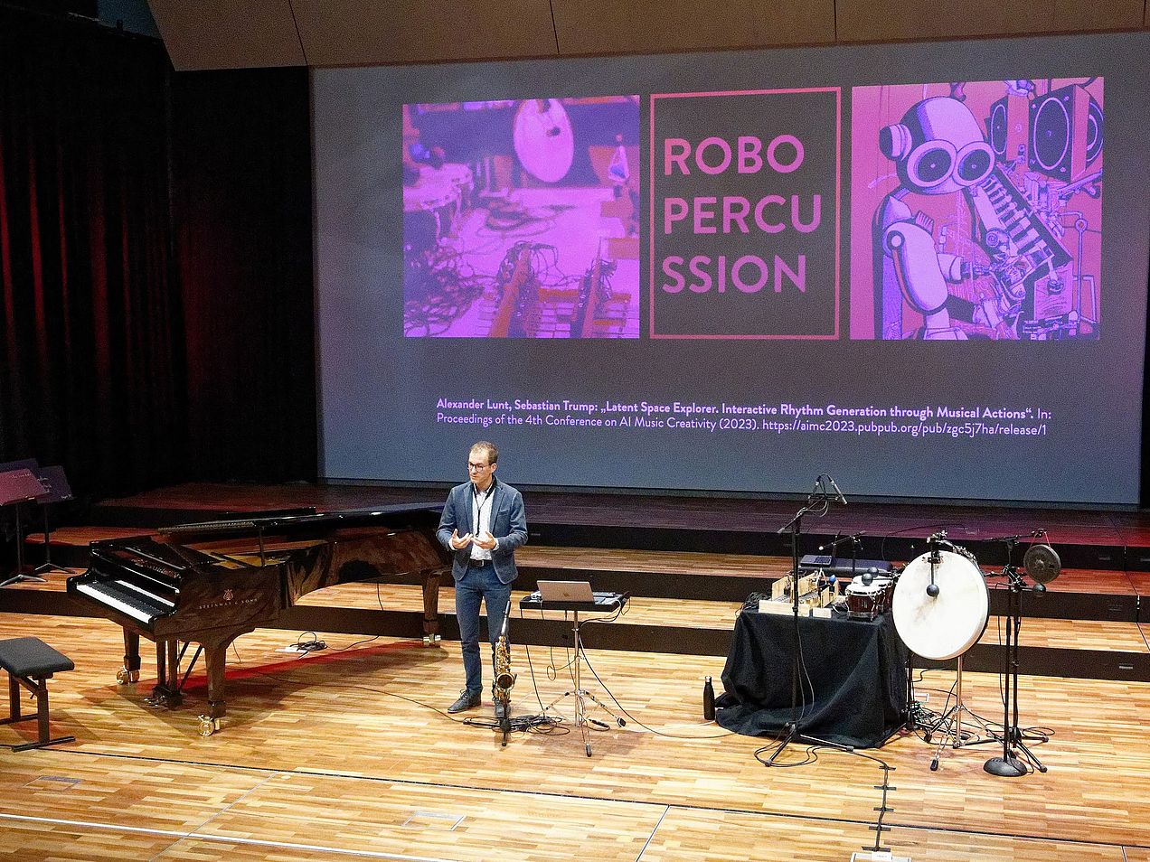 Prof. Dr. Sebastian Trump steht auf der Bühne des Orchestersaals zwischen dem Spirio-Flügel und der RoboPercussions-Band
