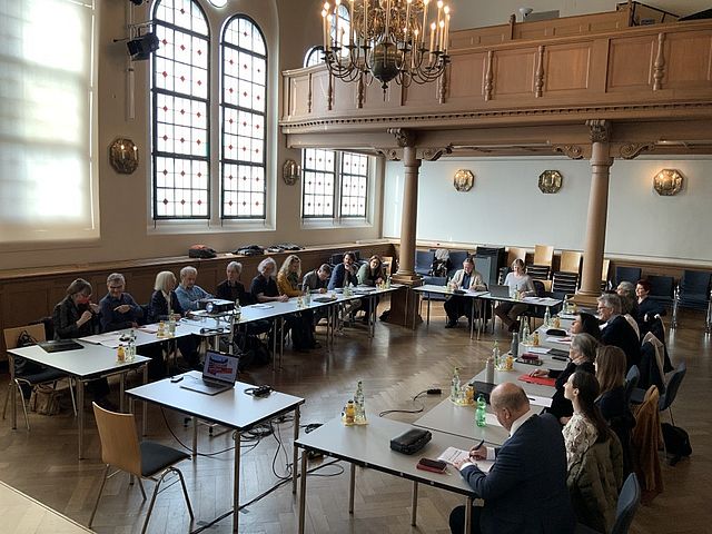 Gruppe sitzt im Kammermusiksaal an Tischen, die in U-Form gestellt sind. Perspektive von der "offenen Seite" des Us. Im Hintergrund ist die Empore des Kammermusiksaals.