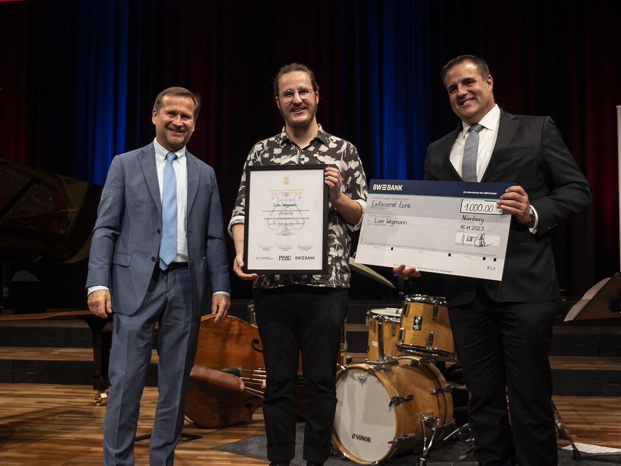Lionel Wegmann steht in der Mitte zwischen Thomas Biller (BW Bank) und Michael Wiesengrund (Piano Haid). Er hält die Steinway-Urkunde und Thomas Biller den 1000€-Scheck in den Händen.