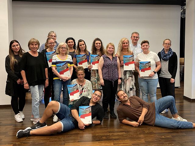 Gruppenbild der 16 Teilnehmenden. Sie stehen in 2 Reihen und halten ihre Zertifikate in den Händen. Zwei Herren liegen auf dem Boden vor der Gruppe. 