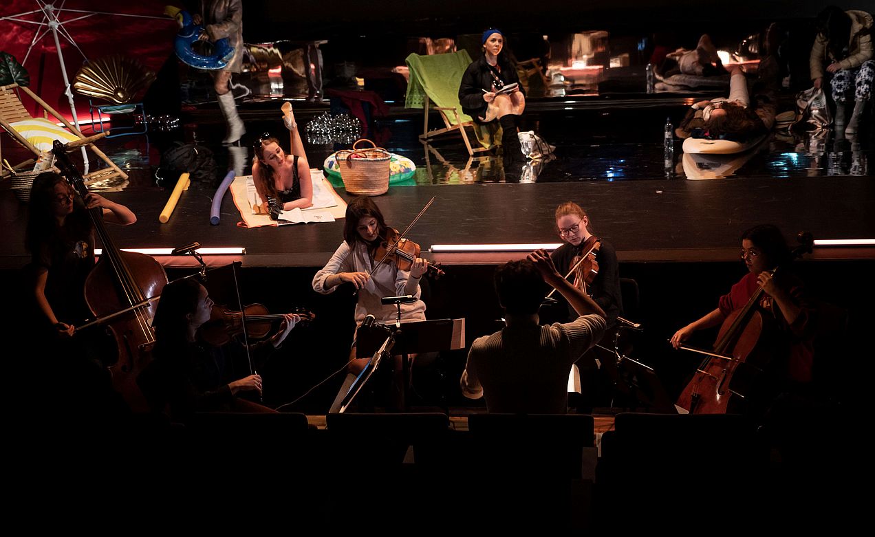 Im Vordergrund befindet sich ein Streichensemble, das vor der Bühne positioniert ist. Auf der Bühne liegt eine Darstellerin auf einer Strandmatte, neben ihr steht eine Strandtasche und ein großer aufblasbarer Schwimmring. Eine weitere Darstellerin singt und sitzt auf eine Liegestuhl. 