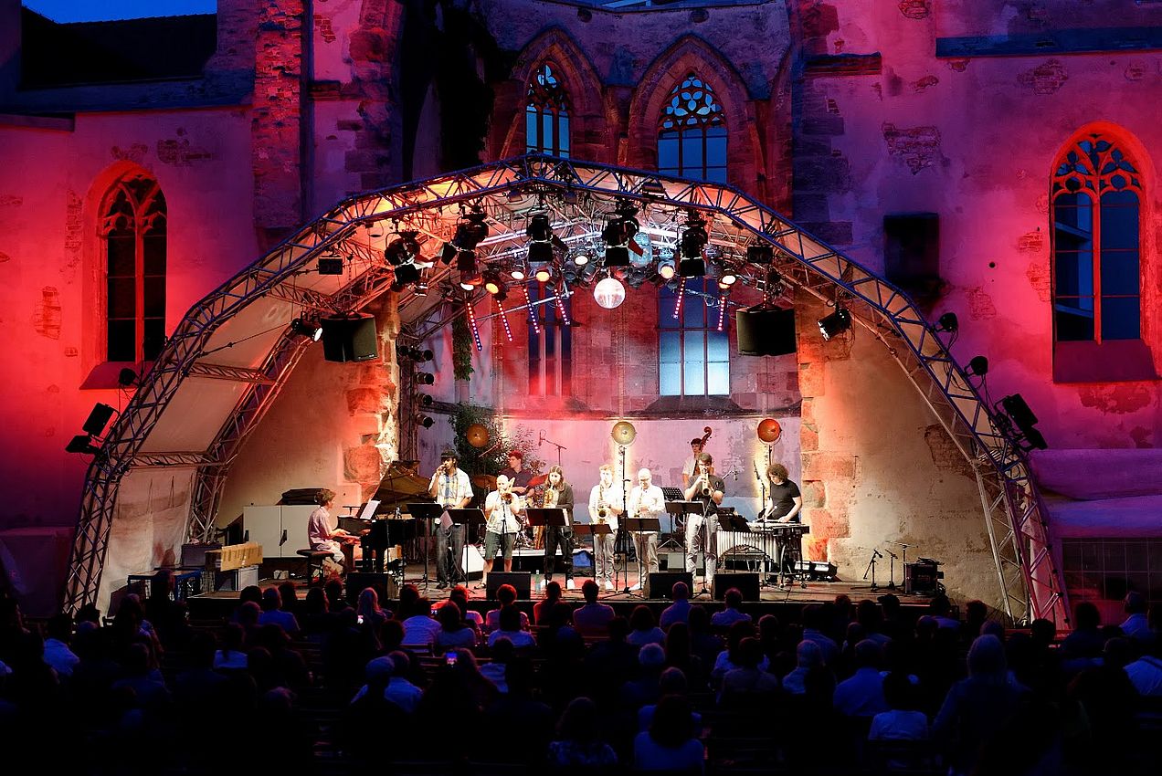 Erleuchtete Bühne vor der Katharinenruine. Mitglieder der Big Band stehen auf der Bühne und spielen Klavier, Saxophon, Trompete, Posaune, Schlagzeug und Bass bzw. singen. 