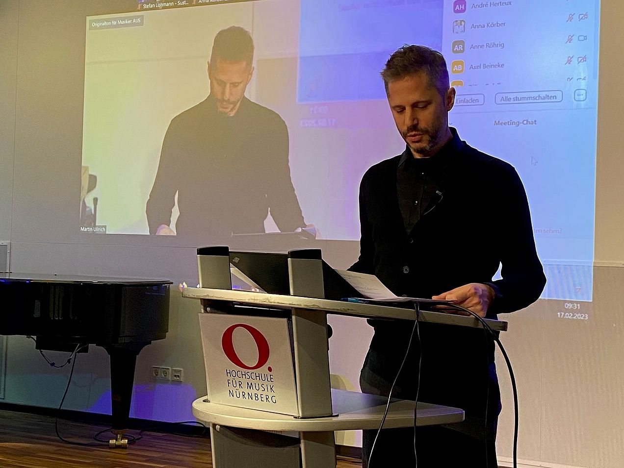 Prof. Rainer Kotzian steht am Redner-Pult, auf dem das Logo der Hochschule für Musik Nürnberg zu sehen ist. 