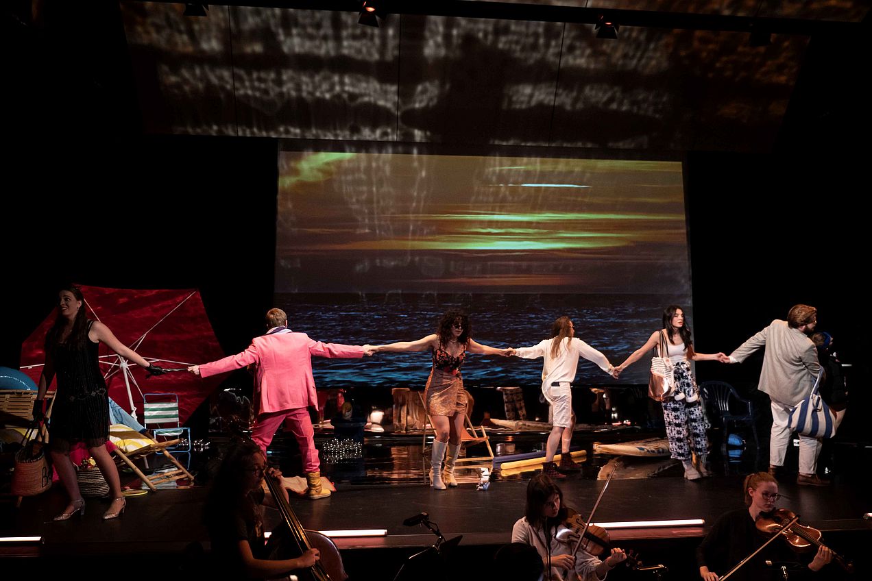 Auf der Bühne halten sich die Darsteller*innen mit ausgestreckten Armen an den Händen. Im Hintergrund stehen Requisiten und an die Wand ist ein Bild von einem Sonnenuntergang über dem Meer angestrahlt. Vor der Bühne sind Mitglieder des Streichensembles zu sehen. 