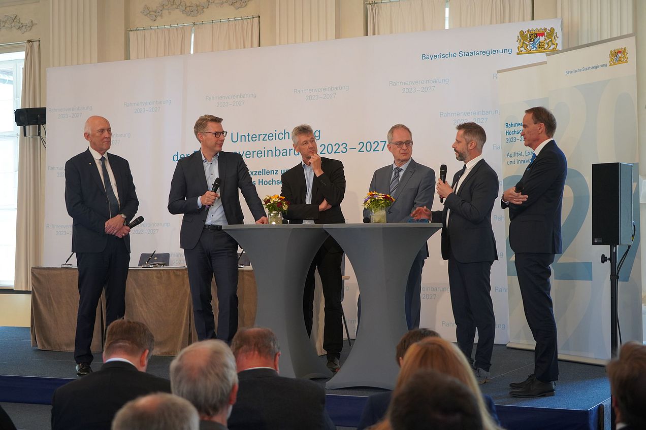 6 Herren stehen im Rahmen der Talkrunde um einen Stehtisch, drei von ihnen halten Mikrofone in den Händen. 