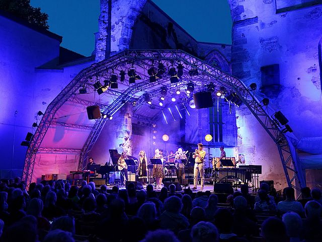 Erleuchtete Bühne vor der Katharinenruine. Mitglieder der Big Band stehen auf der Bühne und spielen Klavier, Trompete, Posaune, Schlagzeug und Bass bzw. singen.