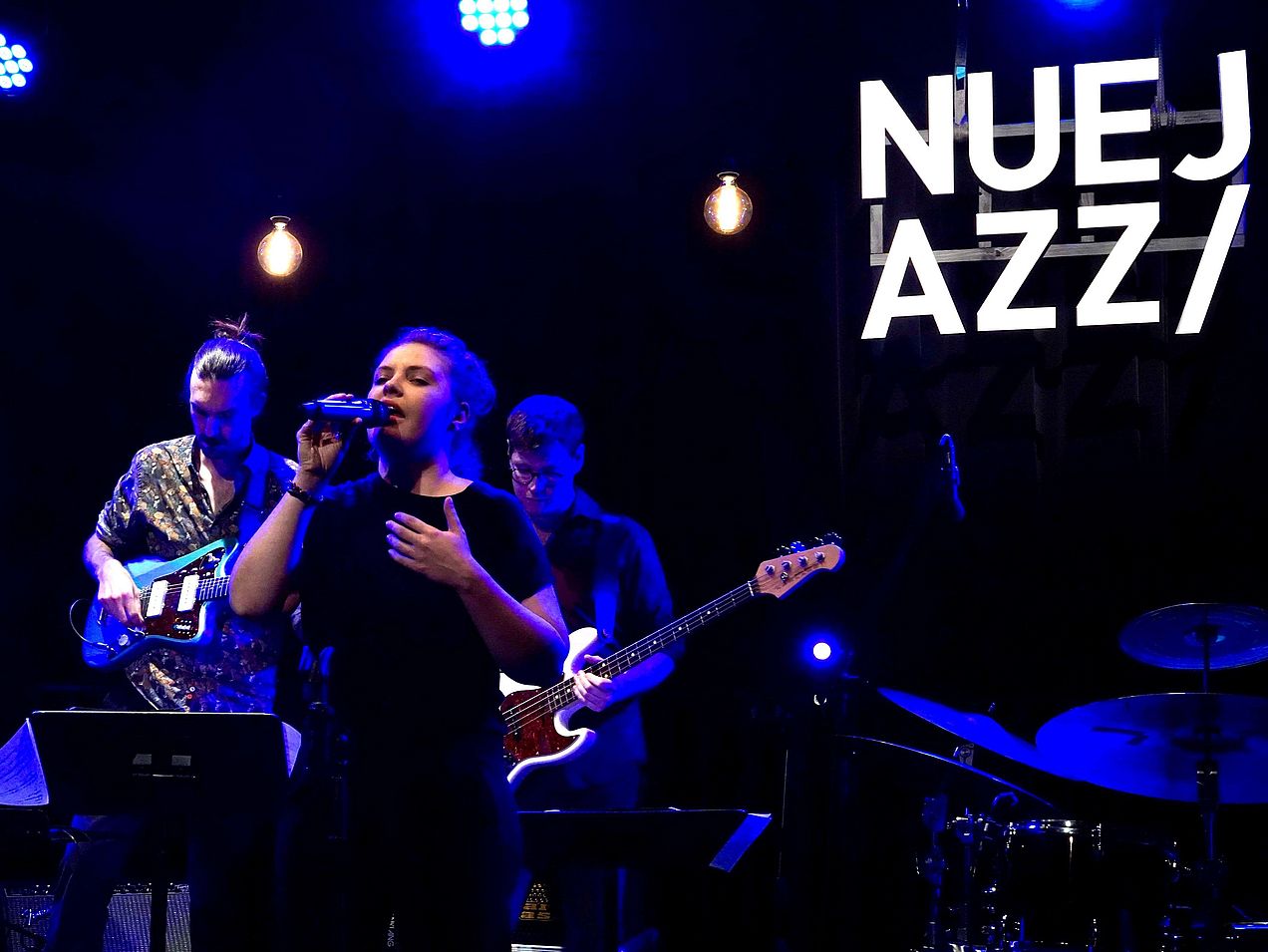 Sängerin Clarissa Forster steht im Vordergrund, hinter ihr befinden sich zwei Gitarristen. An der Wand ist vor schwarzem Hintergrund das weiße Logo des NueJazz Festivals angestrahlt. 