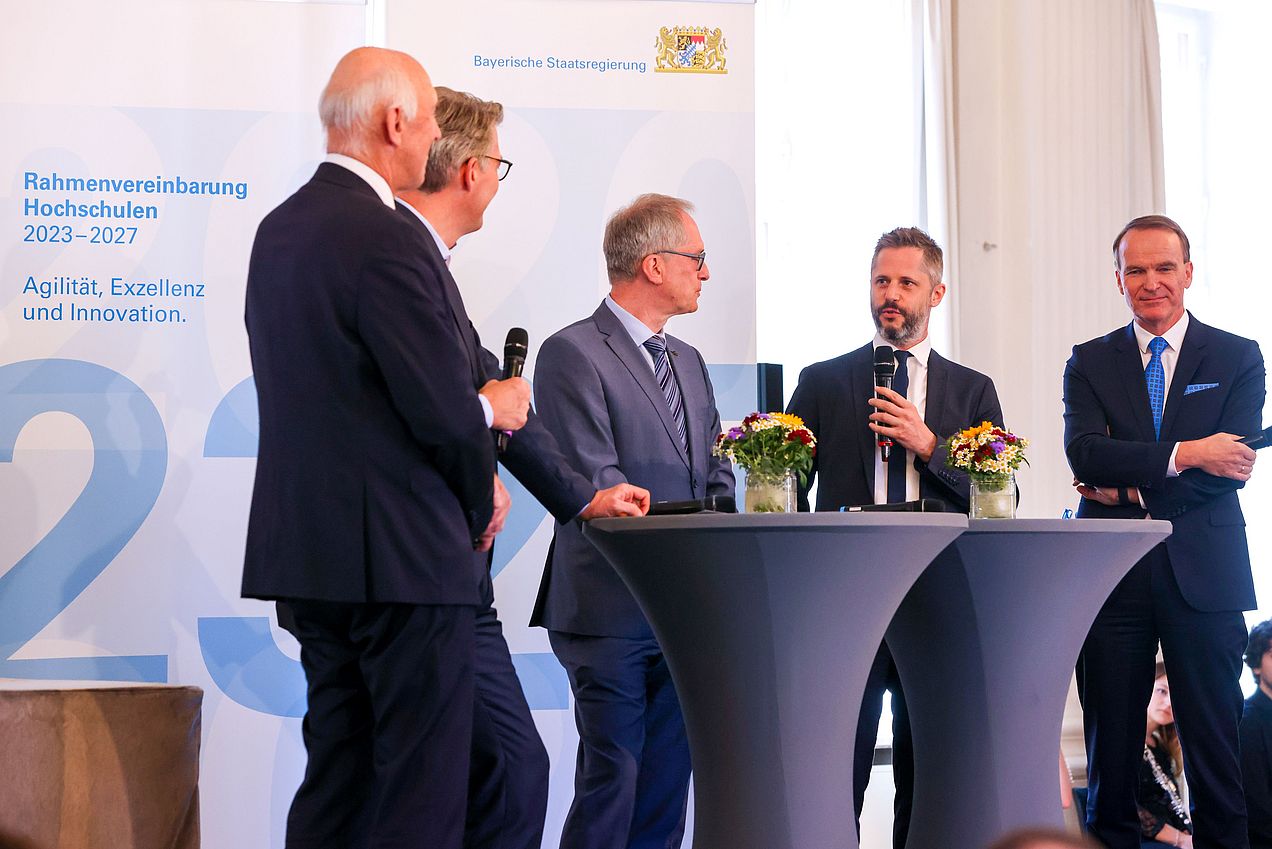 5 Herren stehen im Rahmen der Talkrunde um einen Stehtisch, zwei von ihnen halten Mikrofone in den Händen. 