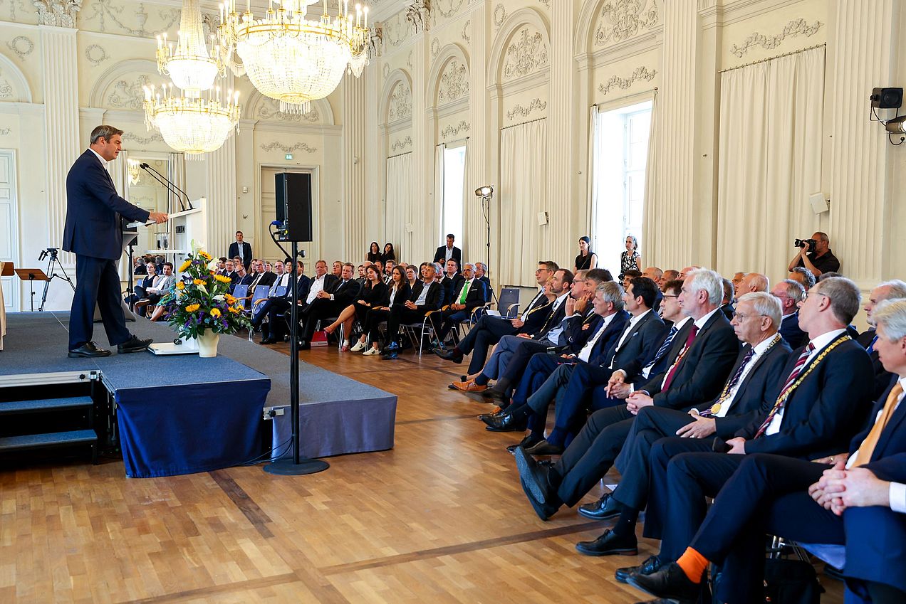 Ministerpräsident Markus Söder steht am Rednerpult auf einer Bühne. Im Publikum sitzen die Präsident*innen der unterzeichnenden Hochschulen und Universitäten. 