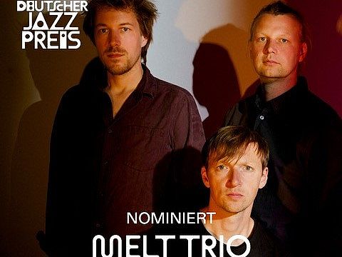 Zwei Herren stehen hinten, ein weiterer sitzt vor ihnen. Logos Deutscher Jazzpreis in der linken oberen Ecke und Text "Preisträger:innen Melttrio" am unteren Bildrand.