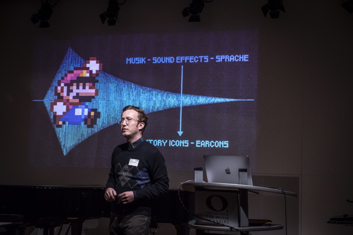 Dr. Pascal Rudolph steht neben dem Rednerpult. im Hintergrund ist die Titelseite seiner Präsentation zur Geschichte der Videospielmusik zu sehen. 
