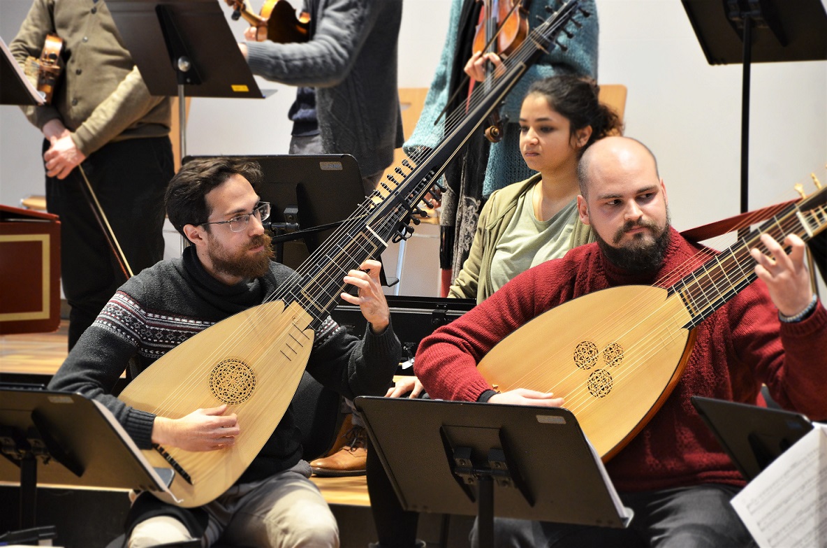 Zwei Männer sitzen mit ihren Lauten nebeneinander. Im Hintergrund befinden sich weitere Musiker*innen mit ihren Instrumenten. 