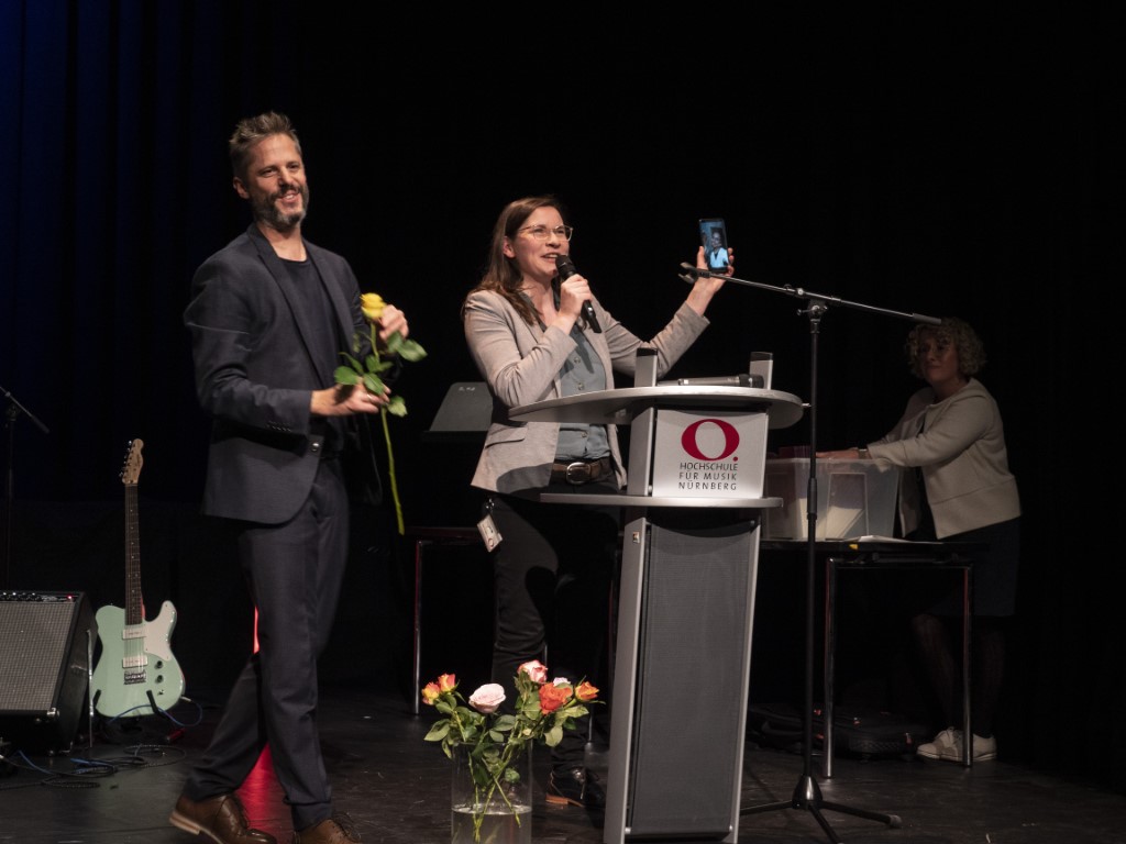 Prof. Rainer Kotzian und Anna Körber stehen auf der Bühne hinter dem Rednerpult. Anna Körber hält ein Handy hoch, über das ein Student via Facetime zugeschaltet ist. 